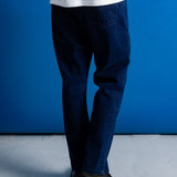 A person shown from the waist down wearing dark blue slim-fit jeans, a white t-shirt, and black boots, standing against a solid blue studio backdrop.