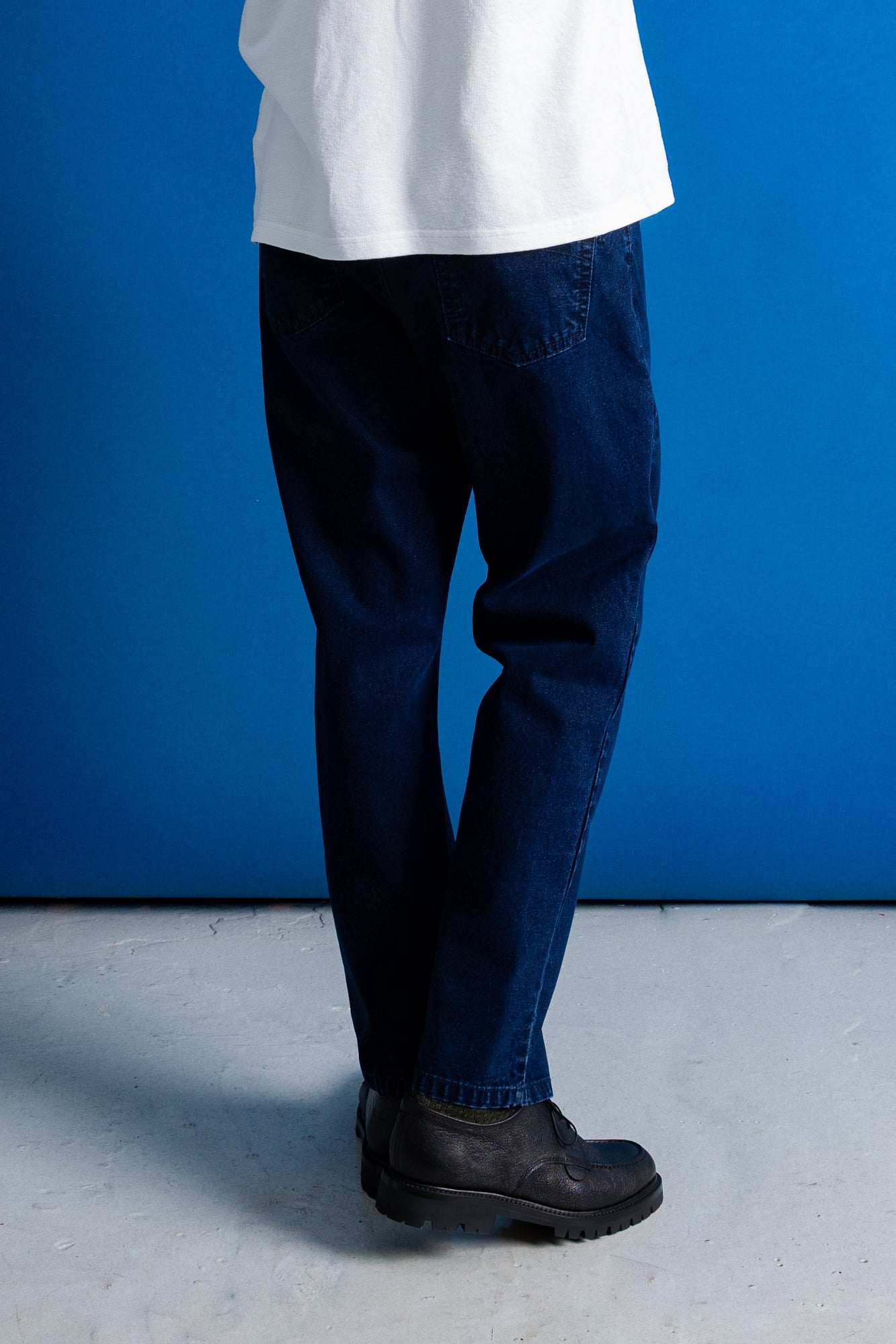 A person shown from the waist down wearing dark blue slim-fit jeans, a white t-shirt, and black boots, standing against a solid blue studio backdrop.