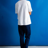 A man viewed from behind wearing a plain white short-sleeve t-shirt, dark blue jeans, and black shoes, standing against a solid blue studio backdrop.