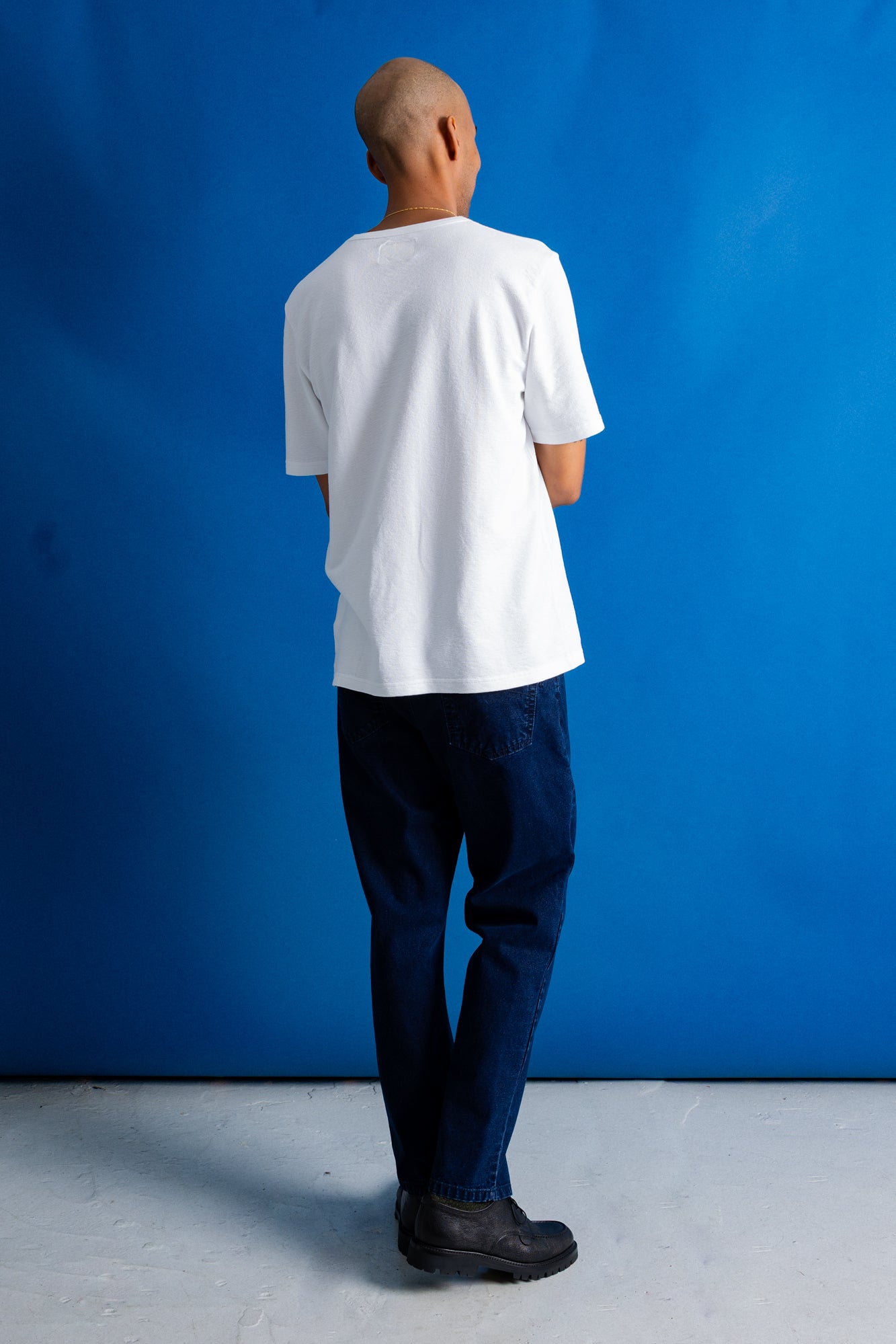 A man viewed from behind wearing a plain white short-sleeve t-shirt, dark blue jeans, and black shoes, standing against a solid blue studio backdrop.