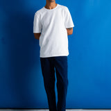 A person from the waist down wearing dark navy blue relaxed-fit trousers, a white t-shirt, and black leather shoes, standing against a vibrant blue wall.