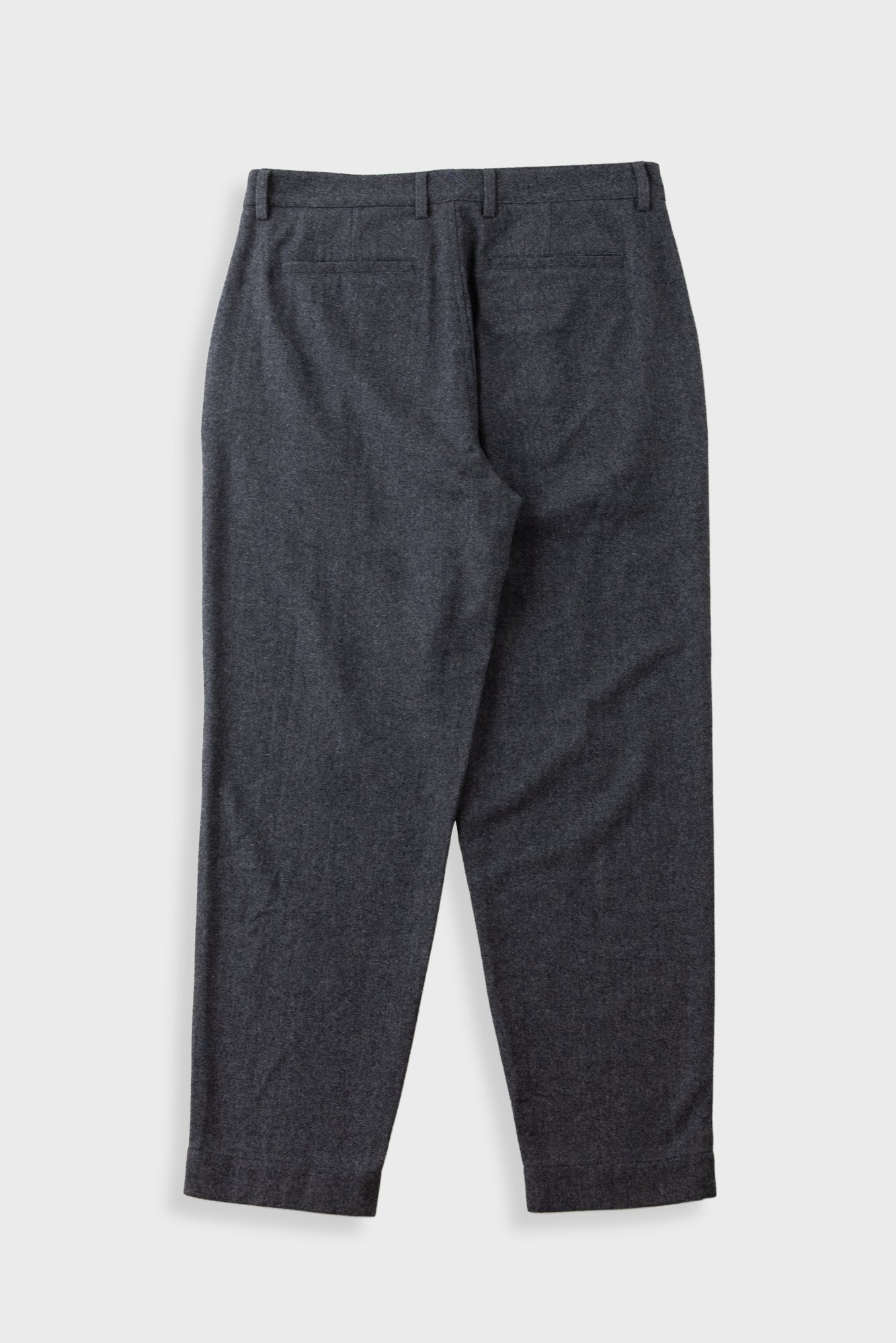 A pair of dark grey pleated trousers laid flat against a solid beige background.