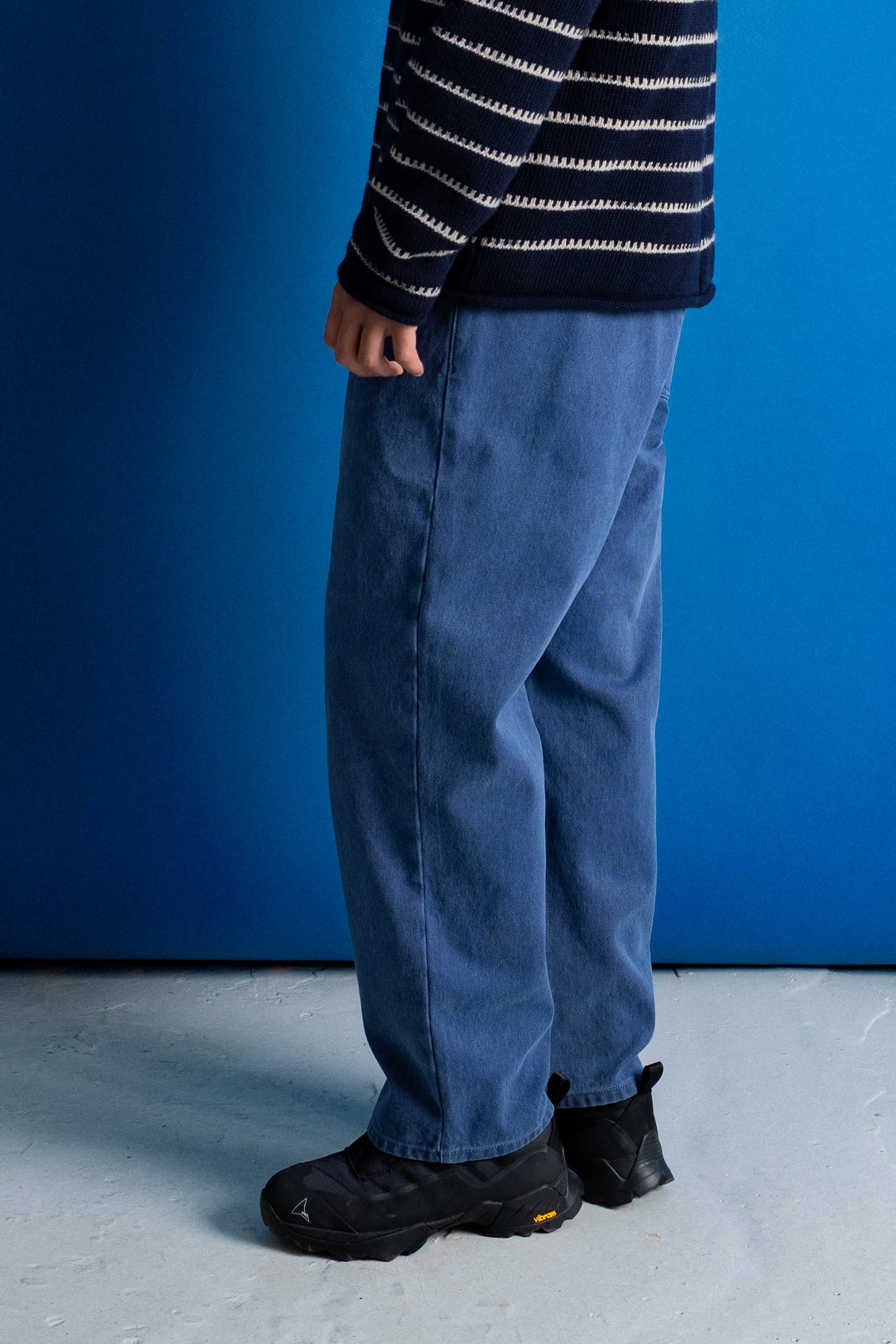A person shown from the waist down wearing loose blue jeans, a navy and white striped sweater, and black boots, standing against a blue wall and light gray floor.