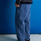 A person shown from the waist down wearing loose blue jeans, a navy and white striped sweater, and black boots, standing against a blue wall and light gray floor.