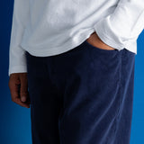 A close-up of a person wearing dark navy corduroy trousers and a white long-sleeve shirt, standing against a solid blue background.