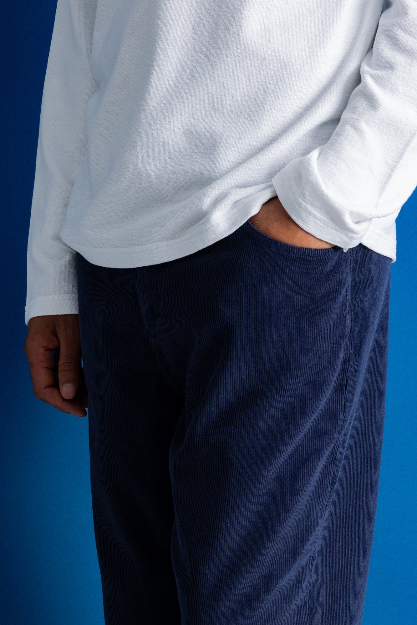 A close-up of a person wearing dark navy corduroy trousers and a white long-sleeve shirt, standing against a solid blue background.