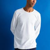A man wearing a plain white long-sleeve crewneck t-shirt and dark navy pants, standing against a solid blue studio background.
