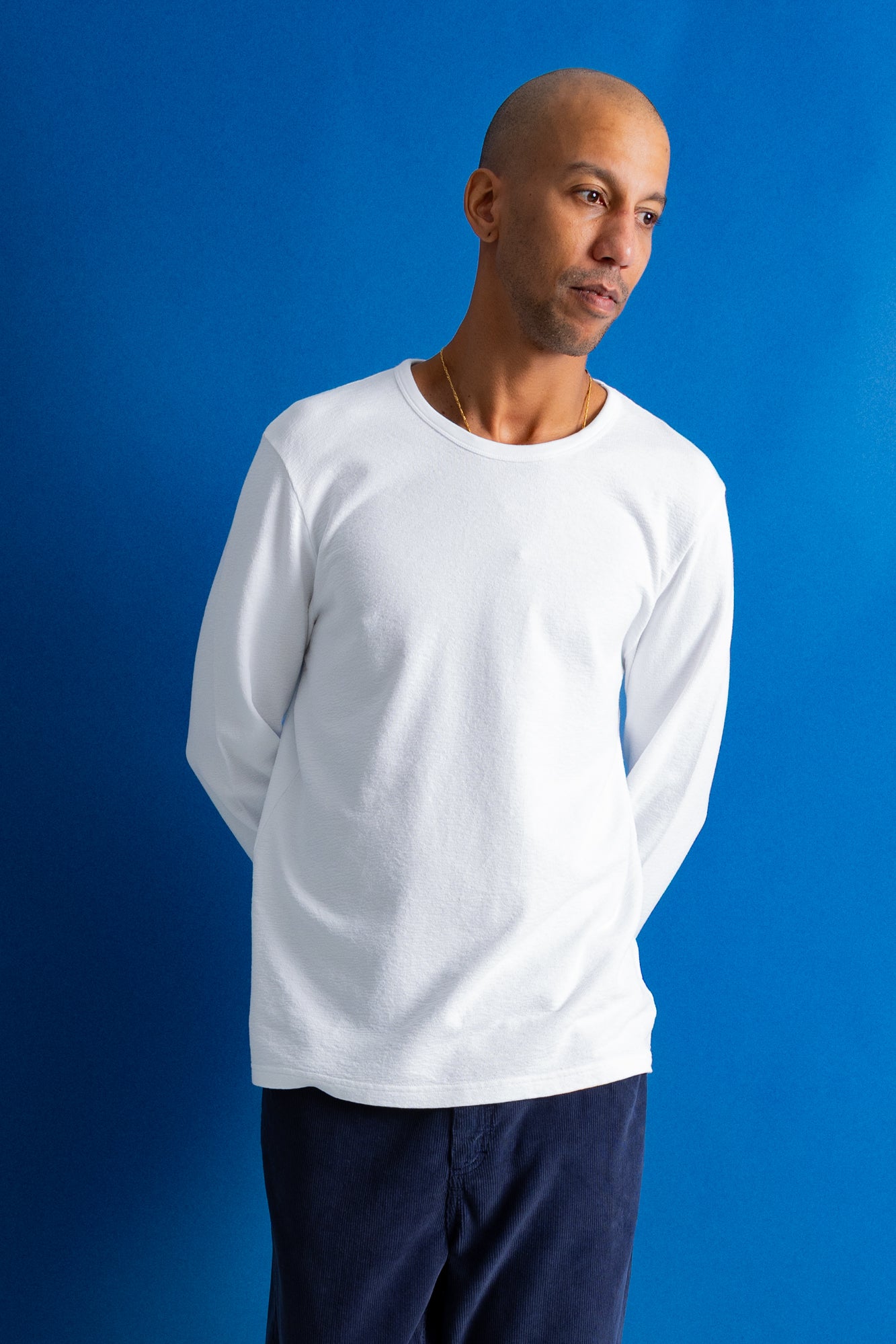 A man wearing a plain white long-sleeve crewneck t-shirt and dark navy pants, standing against a solid blue studio background.