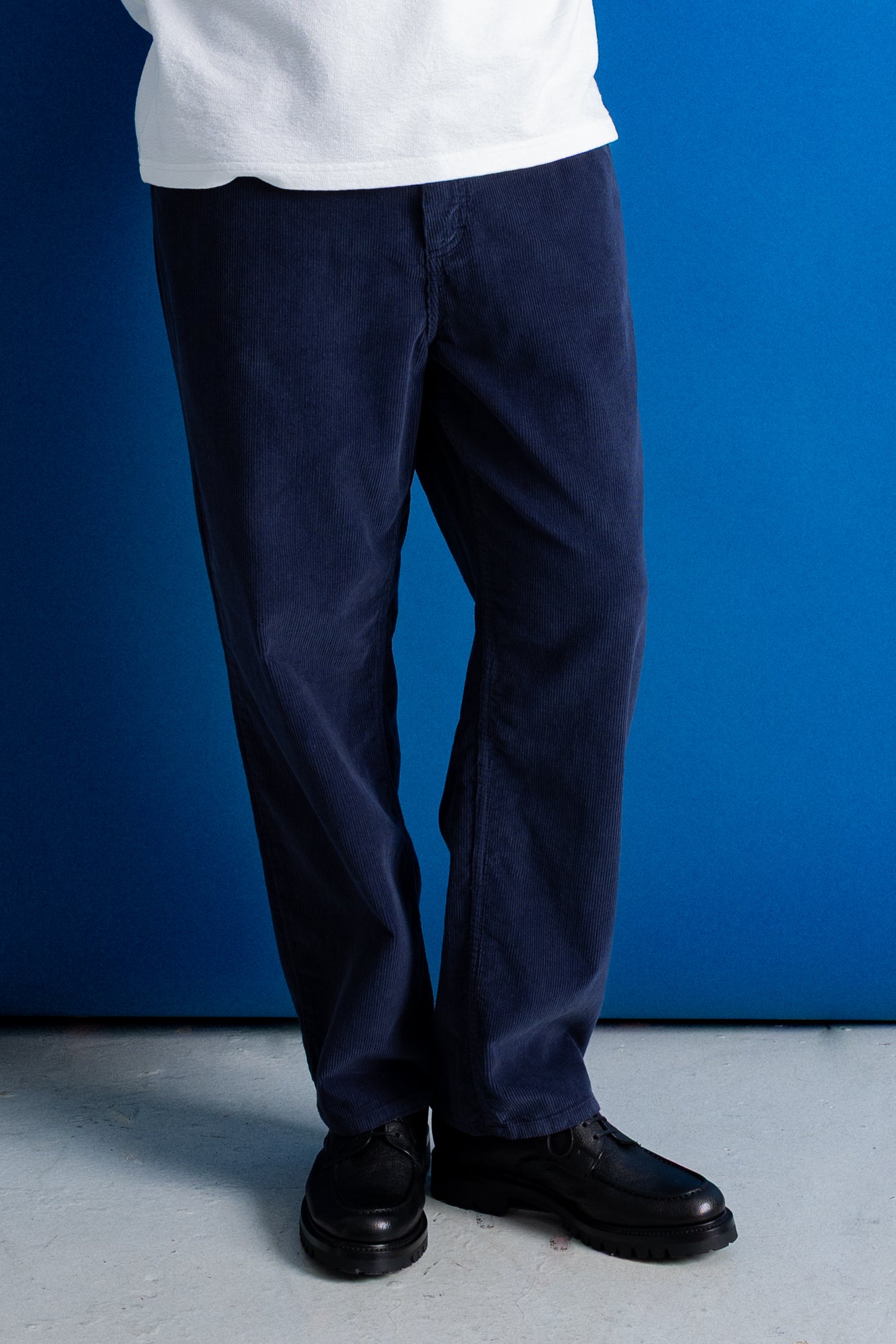 A person wearing dark navy corduroy pants, a white t-shirt, and black leather shoes, standing against a solid blue wall.