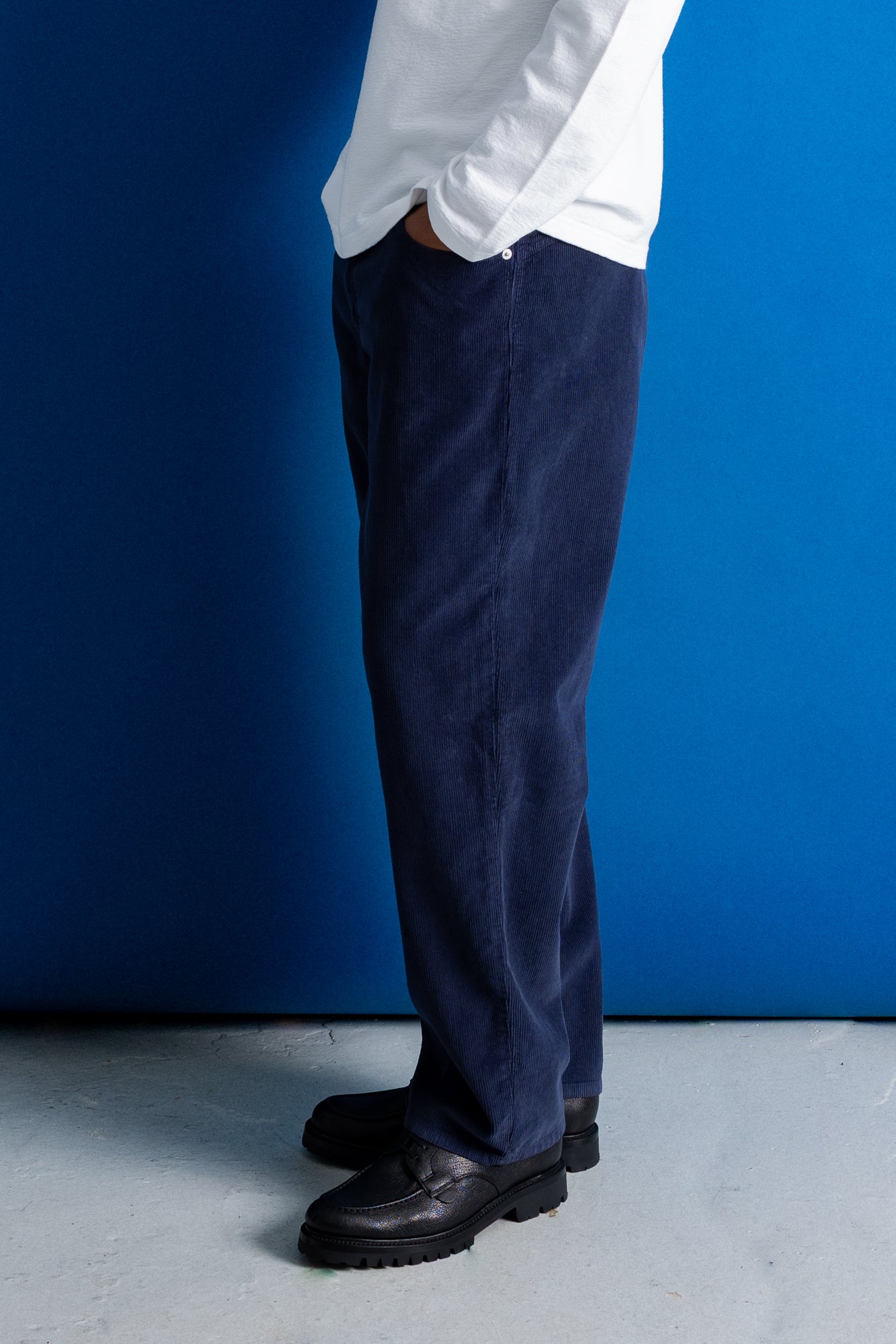 A person shown from the waist down wearing dark navy corduroy trousers, a white shirt, and black leather shoes, standing against a vibrant blue wall.