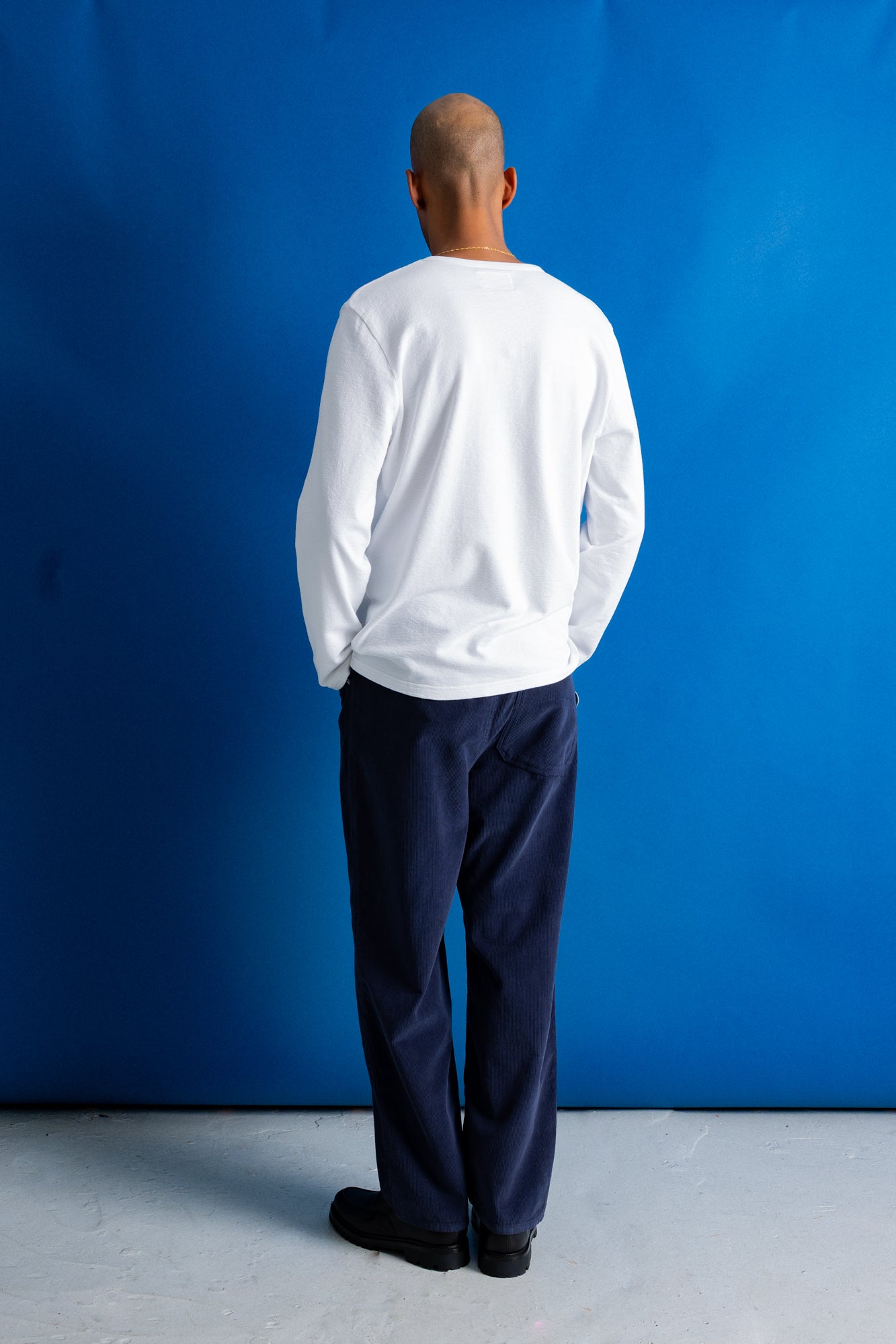 A man viewed from behind wearing a white long-sleeve crewneck shirt and dark navy pants, standing against a vibrant blue backdrop.