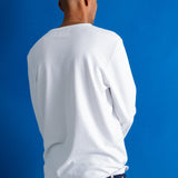 A man viewed from behind wearing a white long-sleeve crewneck shirt and dark navy pants, standing against a solid blue studio background.