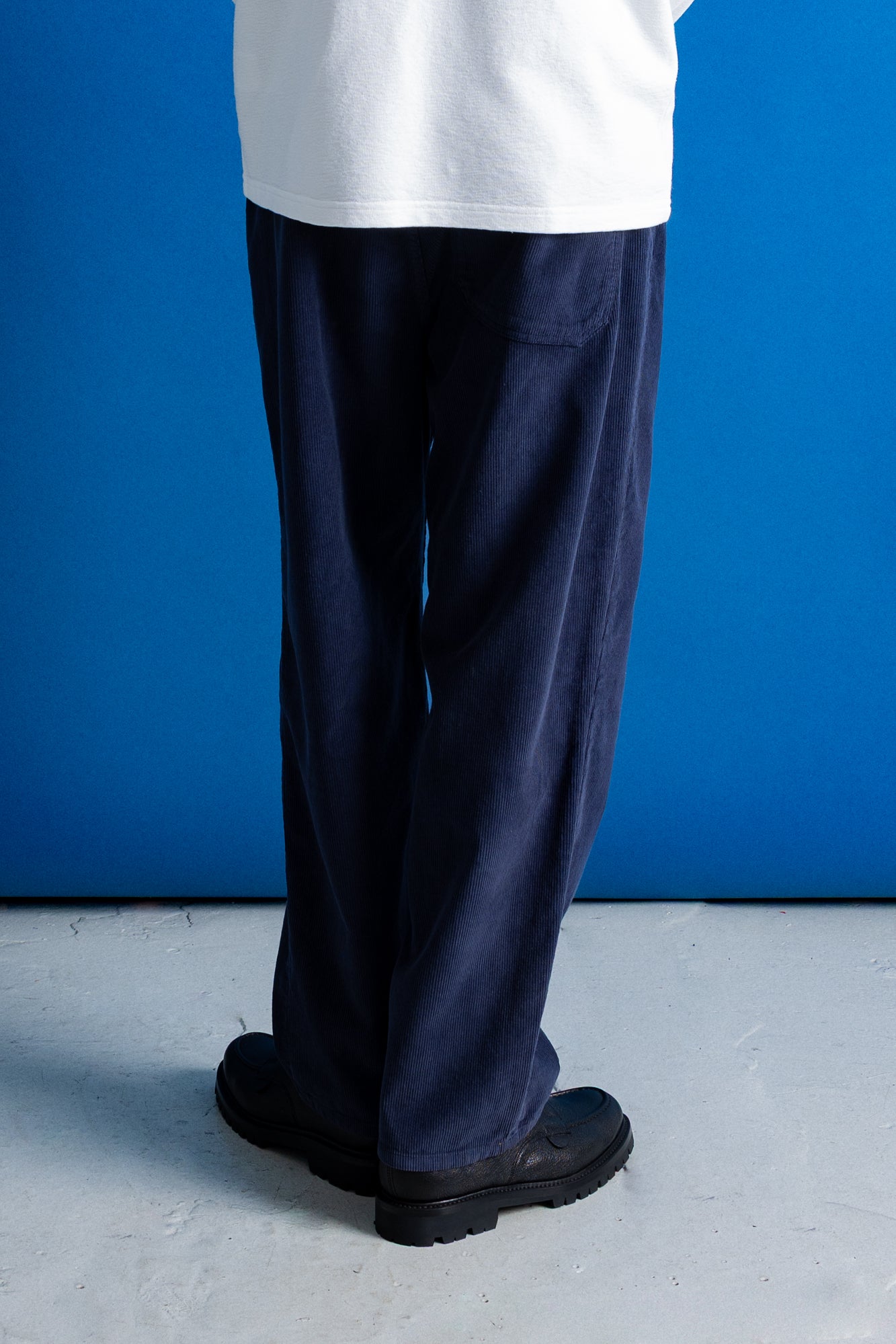 A person shown from the waist down wearing dark navy wide-leg corduroy trousers, a white t-shirt, and black shoes, standing against a vibrant blue wall.