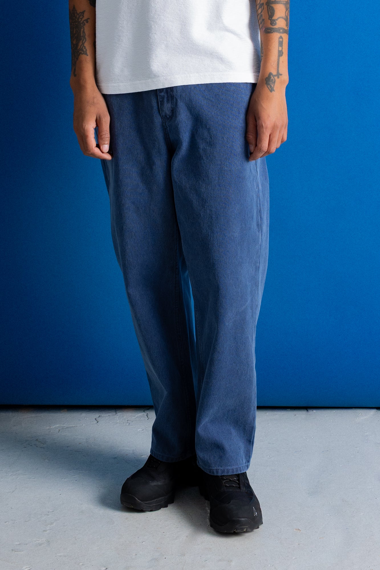 A person wearing loose-fit blue denim pants, a white t-shirt, and black sneakers, standing against a solid blue wall.