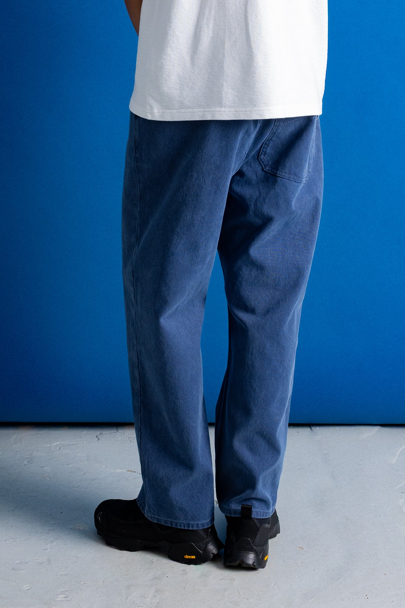 A person shown from the back wearing blue denim jeans, a white t-shirt, and black shoes, standing against a solid blue wall and gray floor.