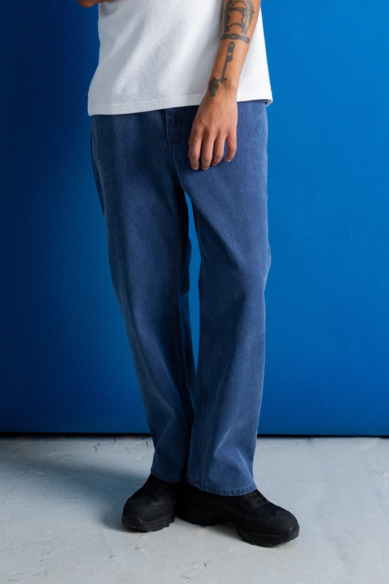 A person wearing loose-fit medium blue denim jeans, a white t-shirt, and black shoes, standing against a solid blue wall.
