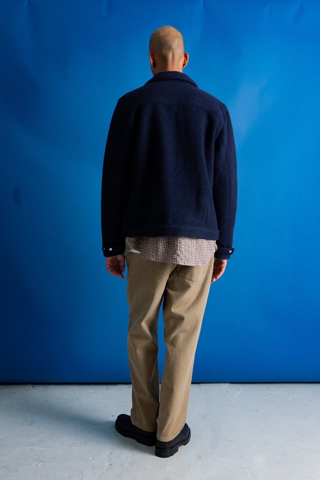 A man viewed from behind wearing a dark navy blue jacket, beige trousers, and black shoes, standing against a bright blue studio backdrop.