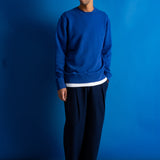 A man wearing a royal blue crewneck sweatshirt with a white hem detail, dark navy pants, and black shoes, standing against a solid blue backdrop.