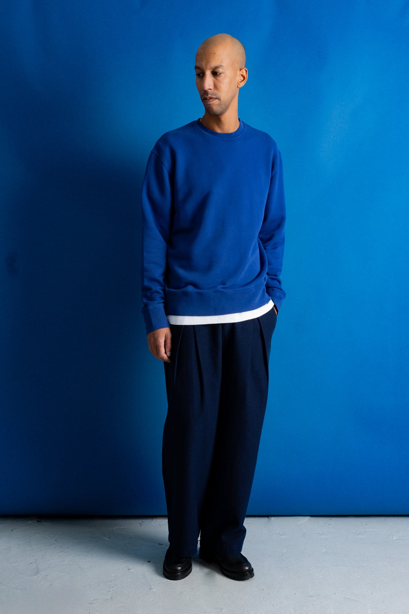 A man wearing a royal blue crewneck sweatshirt with a white hem detail, dark navy pants, and black shoes, standing against a solid blue backdrop.