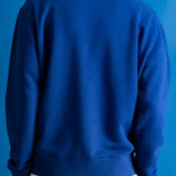 A person viewed from the back wearing a solid royal blue crewneck sweatshirt over a white undershirt, set against a solid blue background.