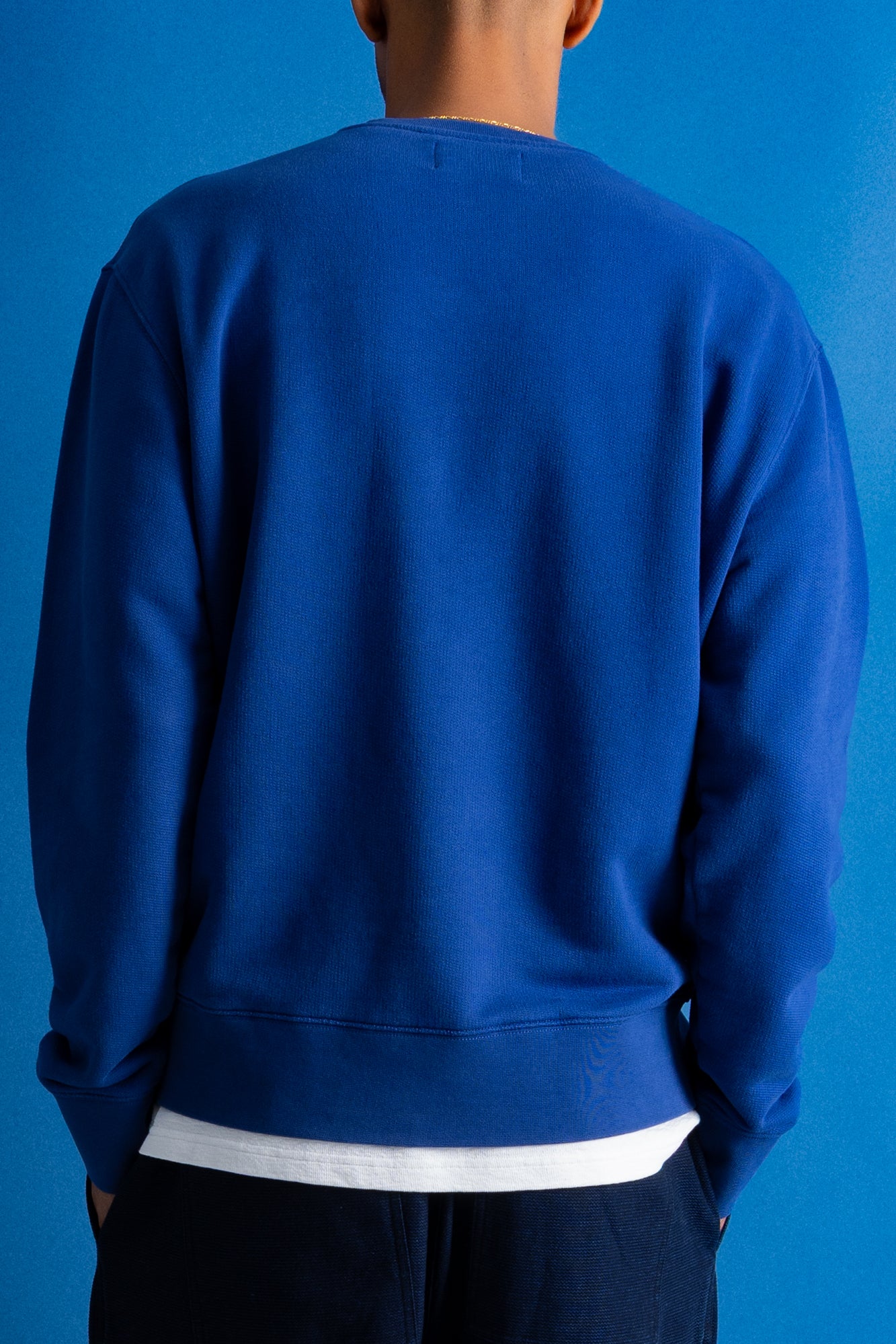 A person viewed from the back wearing a solid royal blue crewneck sweatshirt over a white undershirt, set against a solid blue background.