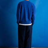 A man viewed from behind wearing a royal blue crewneck sweatshirt, dark navy wide-leg trousers, and black shoes, standing against a vibrant blue studio backdrop.