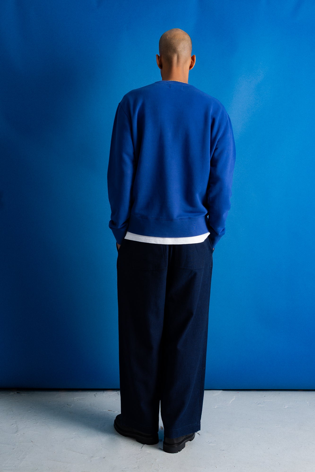 A man viewed from behind wearing a royal blue crewneck sweatshirt, dark navy wide-leg trousers, and black shoes, standing against a vibrant blue studio backdrop.