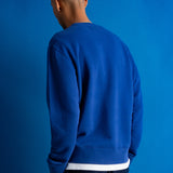 A man viewed from behind wearing a royal blue crewneck sweatshirt with a contrasting white hem, set against a solid blue studio background.