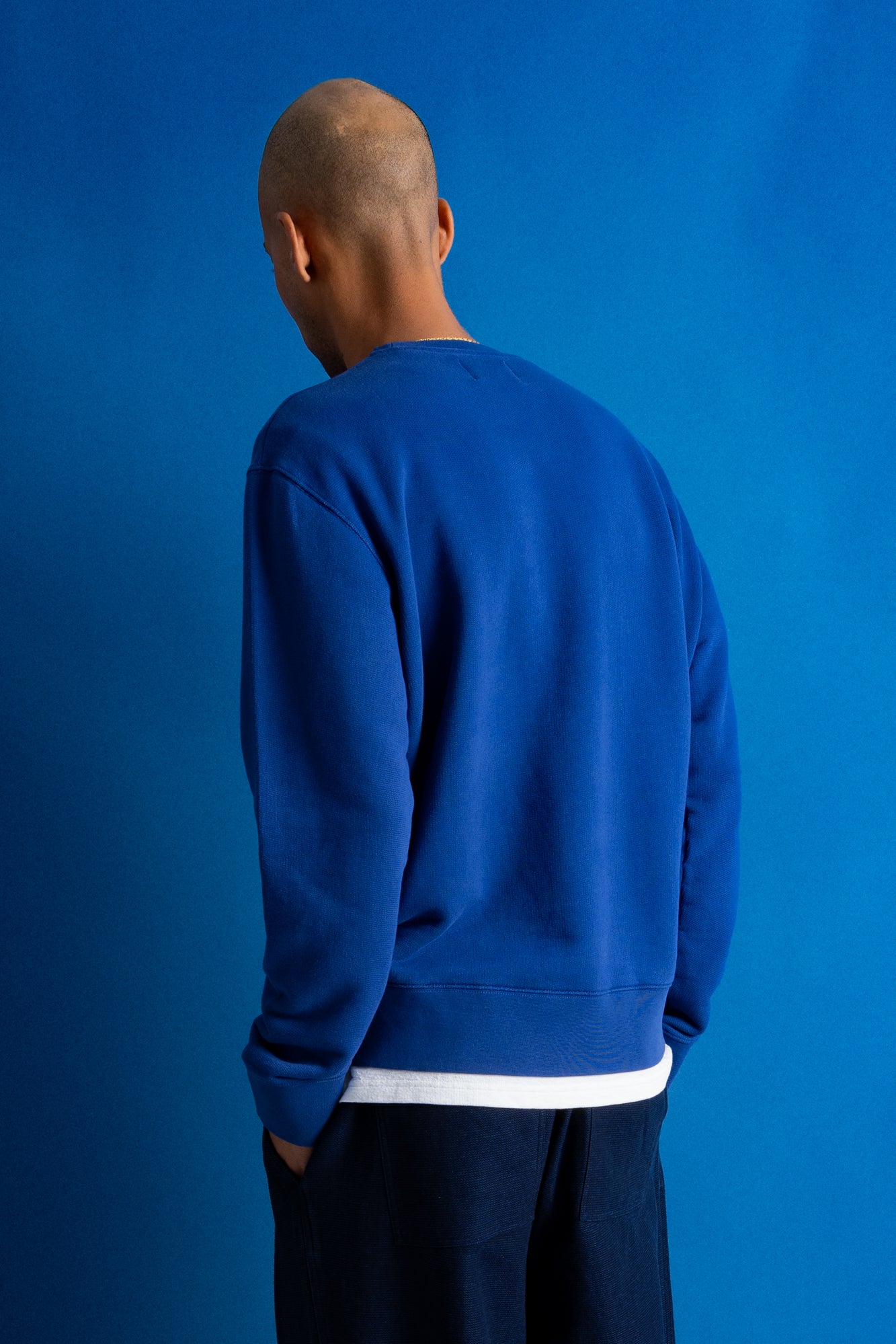 A man viewed from behind wearing a royal blue crewneck sweatshirt with a contrasting white hem, set against a solid blue studio background.