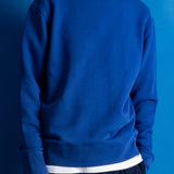 A man wearing a royal blue crewneck sweatshirt with a contrasting white hem, paired with dark trousers, set against a solid blue background.