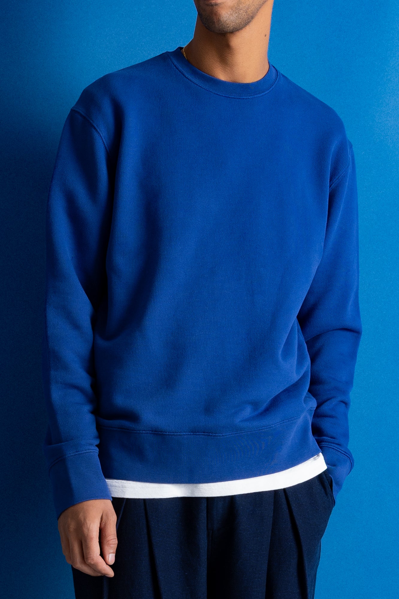 A man wearing a royal blue crewneck sweatshirt with a contrasting white hem, paired with dark trousers, set against a solid blue background.