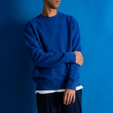 A man wearing a royal blue crewneck sweatshirt and dark navy pants, standing against a solid blue studio background.