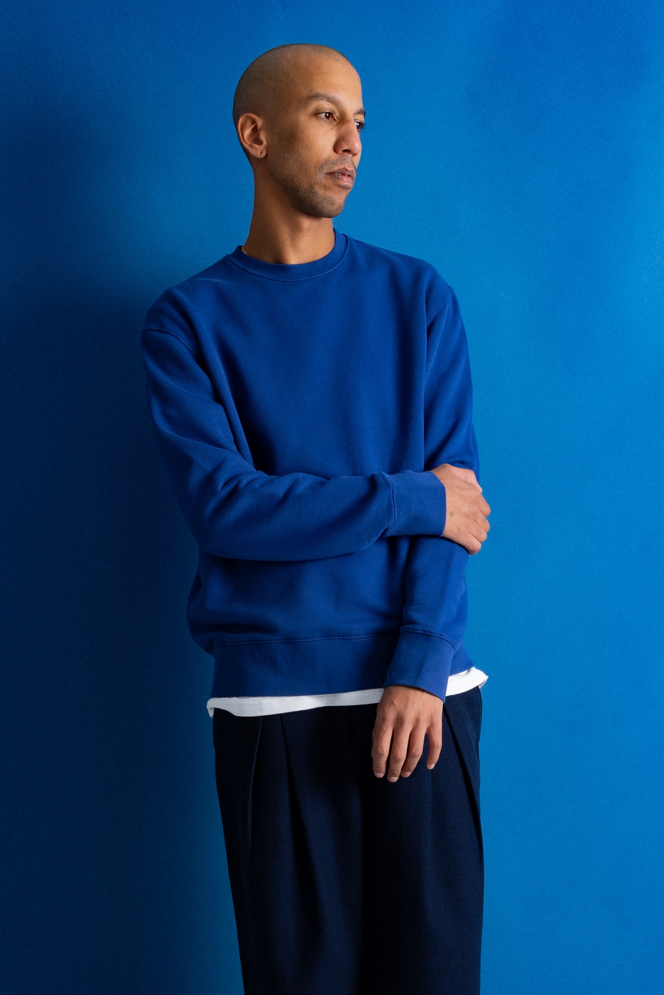 A man wearing a royal blue crewneck sweatshirt and dark navy pants, standing against a solid blue studio background.