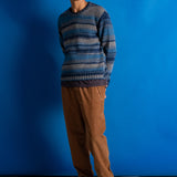 A man wearing a blue and gray striped knit sweater, brown pants, and black shoes, standing against a solid blue studio backdrop.