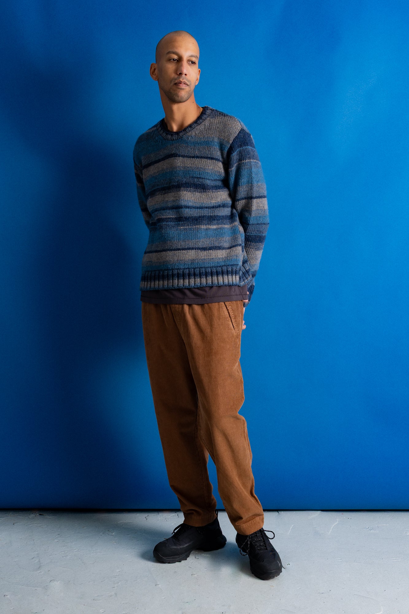 A man wearing a blue and gray striped knit sweater, brown pants, and black shoes, standing against a solid blue studio backdrop.