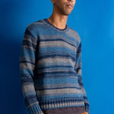 A man wearing a blue and gray striped knit sweater and brown pants, standing against a solid blue background.