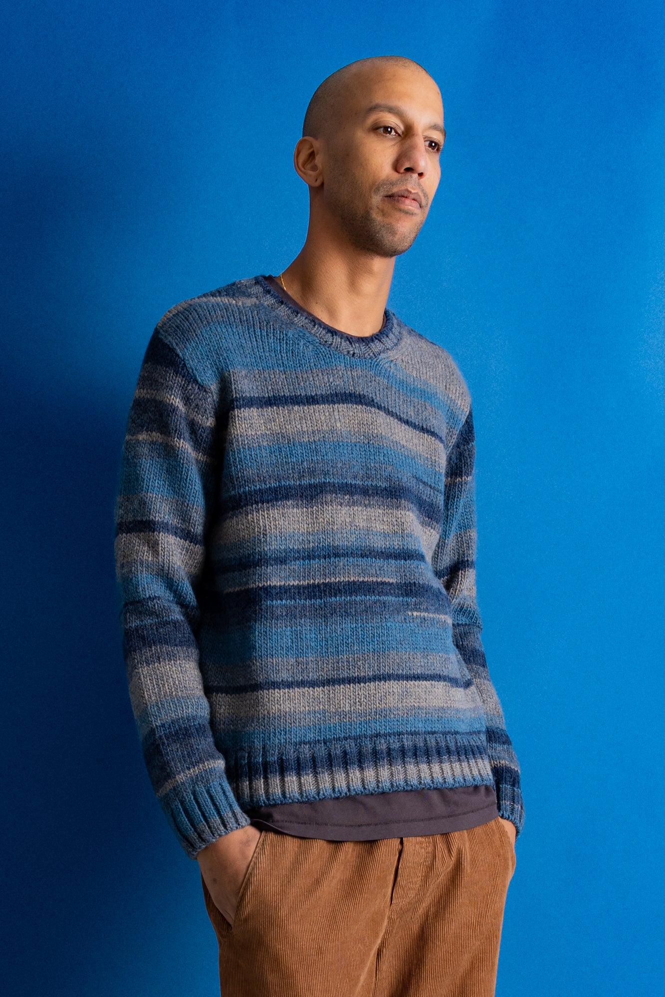 A man wearing a blue and gray striped knit sweater and brown pants, standing against a solid blue background.
