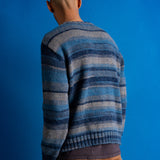 A man viewed from behind wearing a blue and gray striped knit sweater and brown pants, standing against a solid blue studio background.