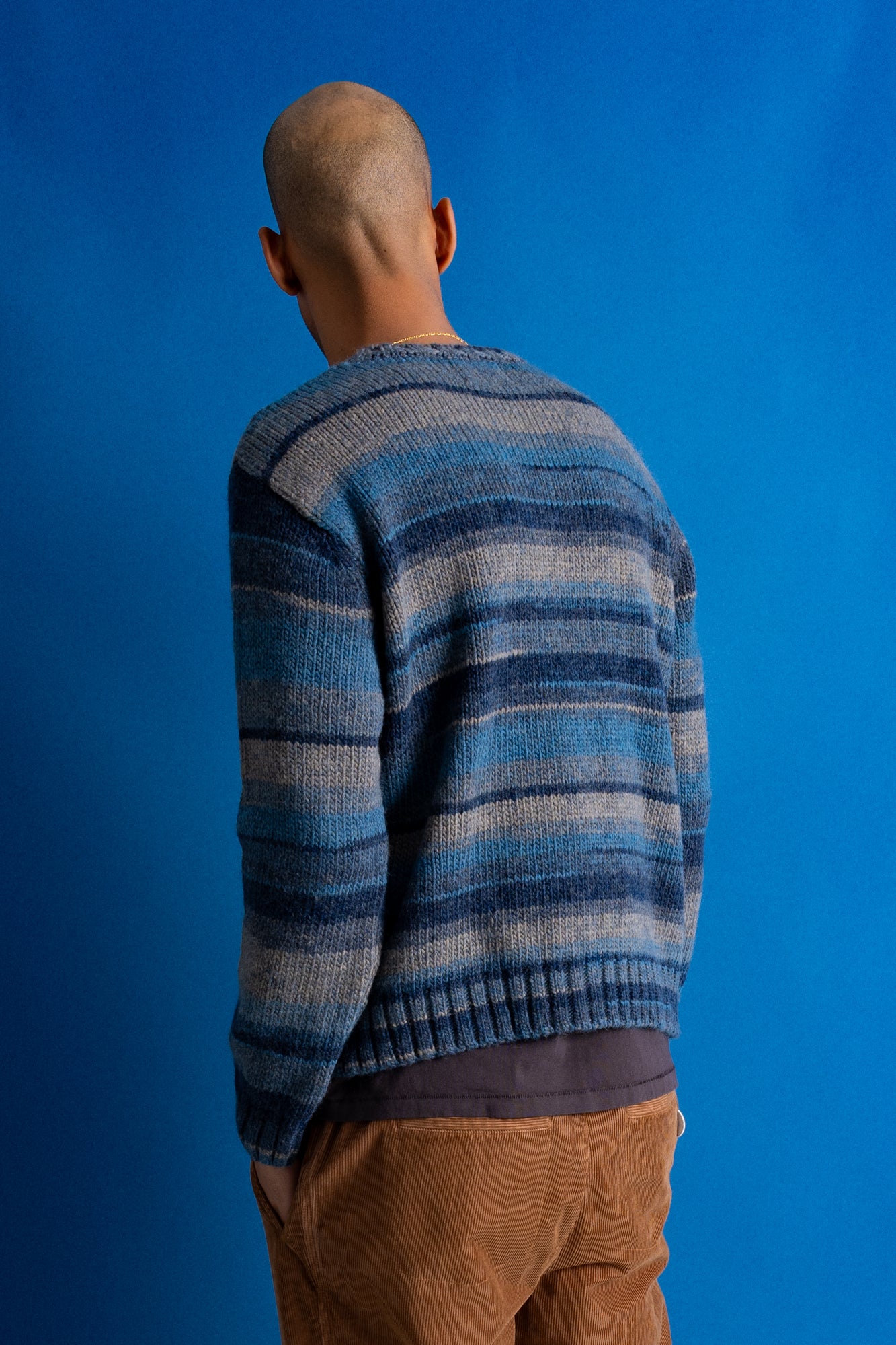 A man viewed from behind wearing a blue and gray striped knit sweater and brown pants, standing against a solid blue studio background.