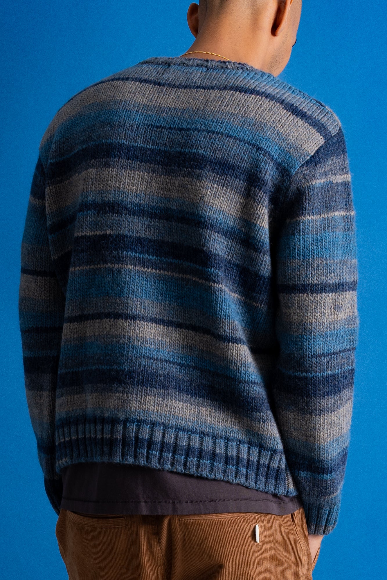 A person shown from the back wearing a blue and gray striped knit sweater, paired with brown pants, against a solid blue background.