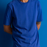 A man wearing a vibrant royal blue crewneck t-shirt, shown from the chest down against a solid blue background.
