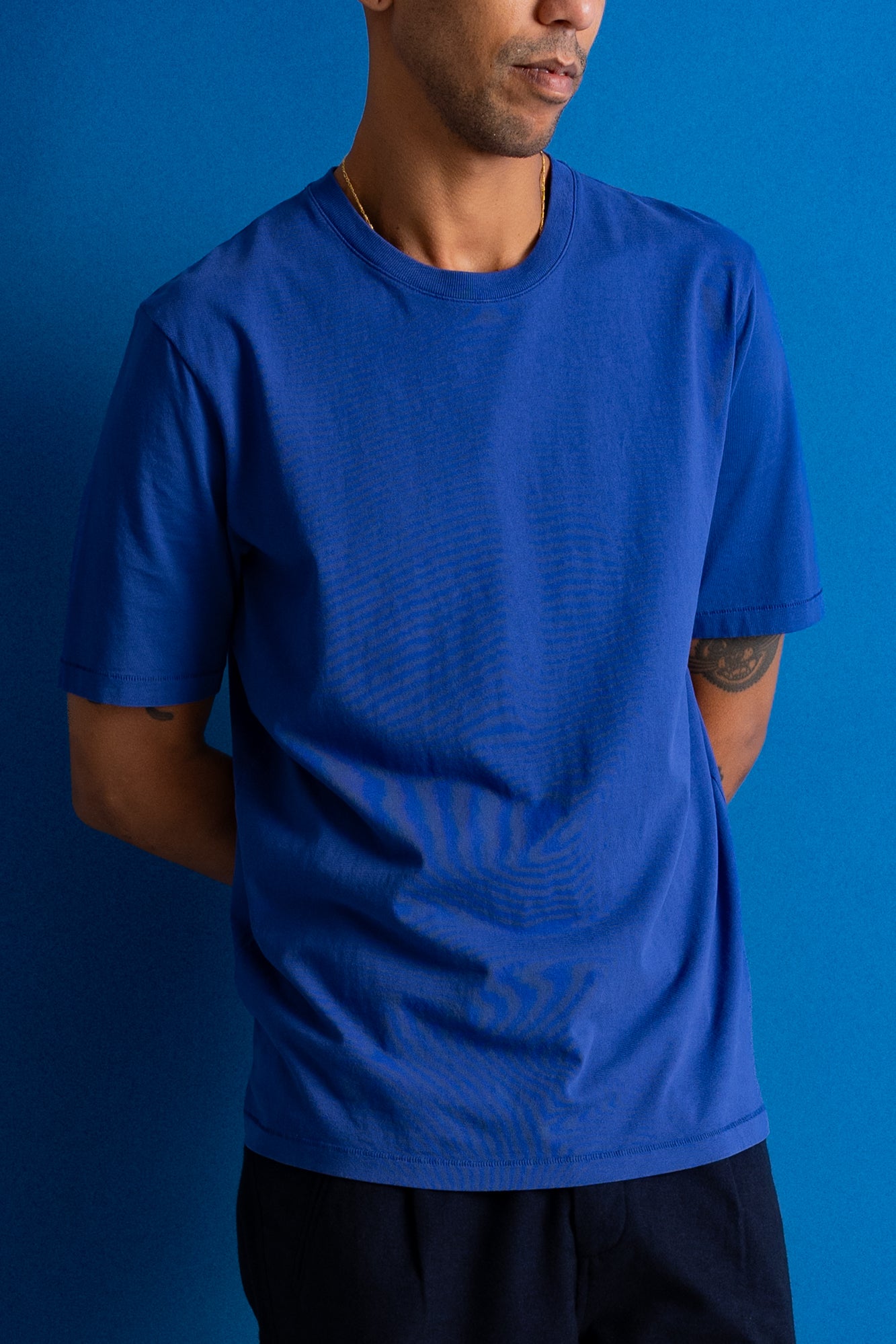 A man wearing a vibrant royal blue crewneck t-shirt, shown from the chest down against a solid blue background.