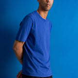 A man wearing a solid royal blue short-sleeve crewneck t-shirt and dark pants, standing against a solid blue studio backdrop.
