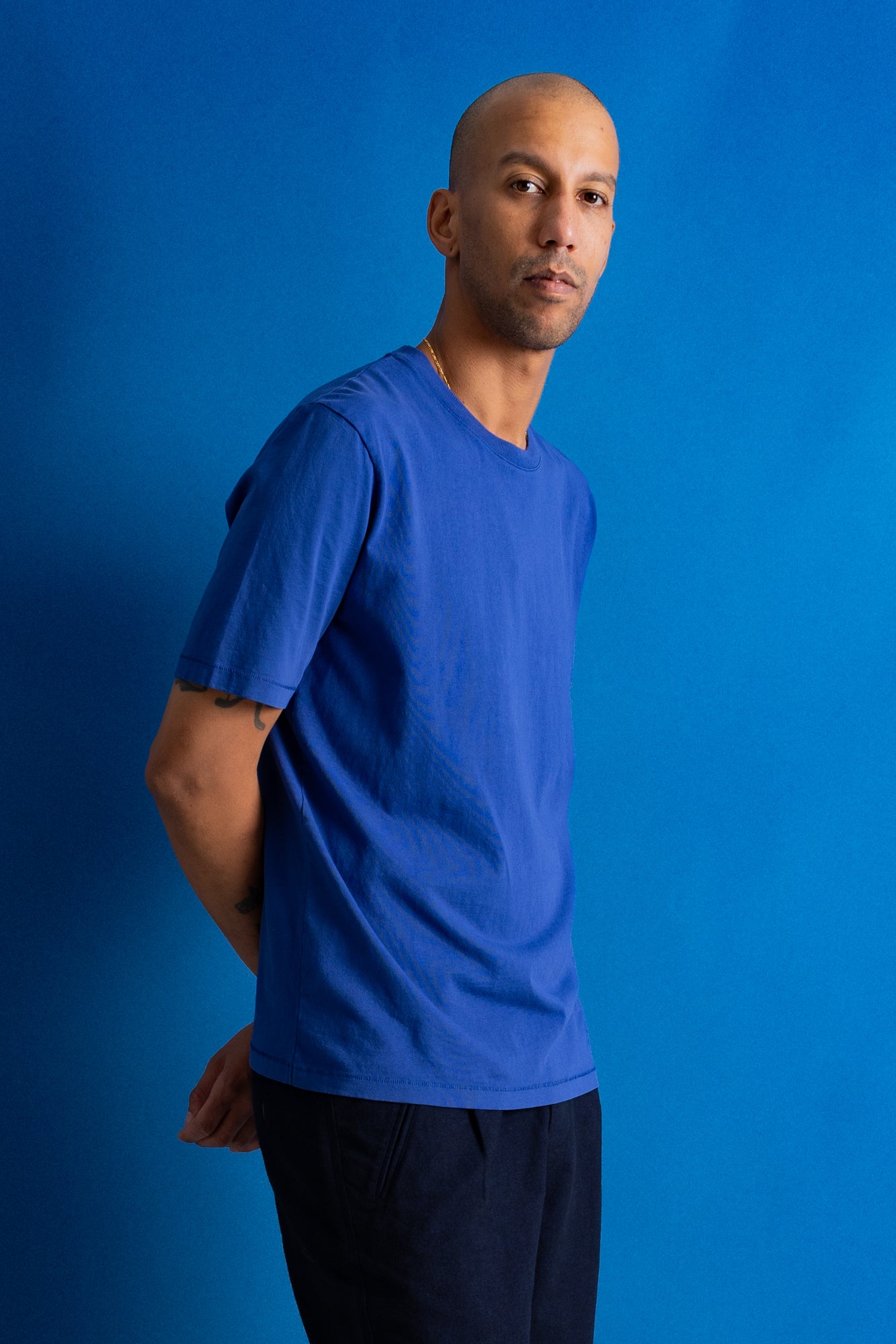 A man wearing a solid royal blue short-sleeve crewneck t-shirt and dark pants, standing against a solid blue studio backdrop.