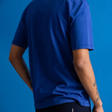 A man wearing a royal blue short-sleeve t-shirt, shown from a rear three-quarter angle against a solid blue studio background.