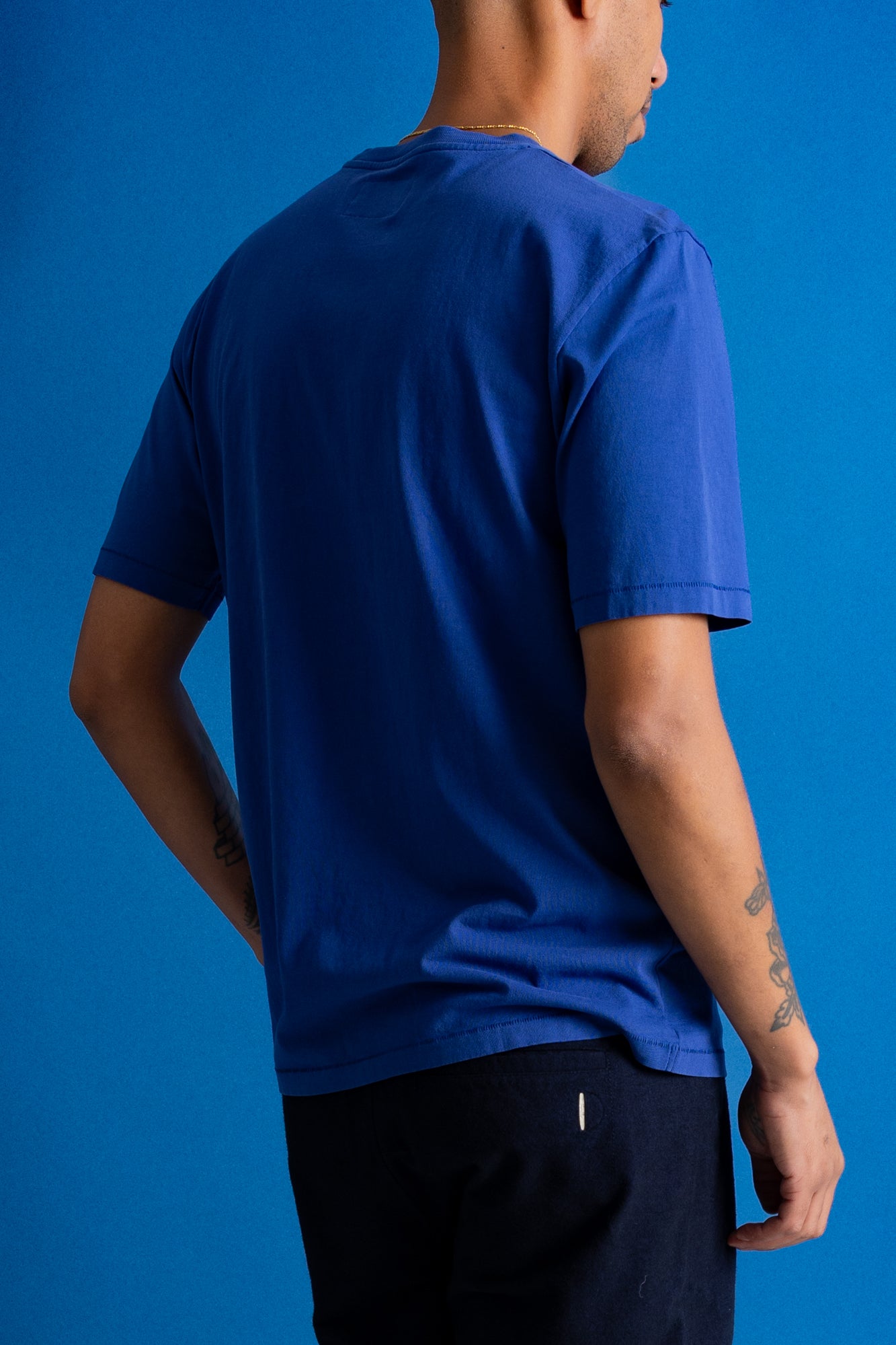 A man wearing a royal blue short-sleeve t-shirt, shown from a rear three-quarter angle against a solid blue studio background.