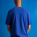 A man viewed from behind wearing a royal blue short-sleeve t-shirt and dark pants, standing against a solid blue studio background.