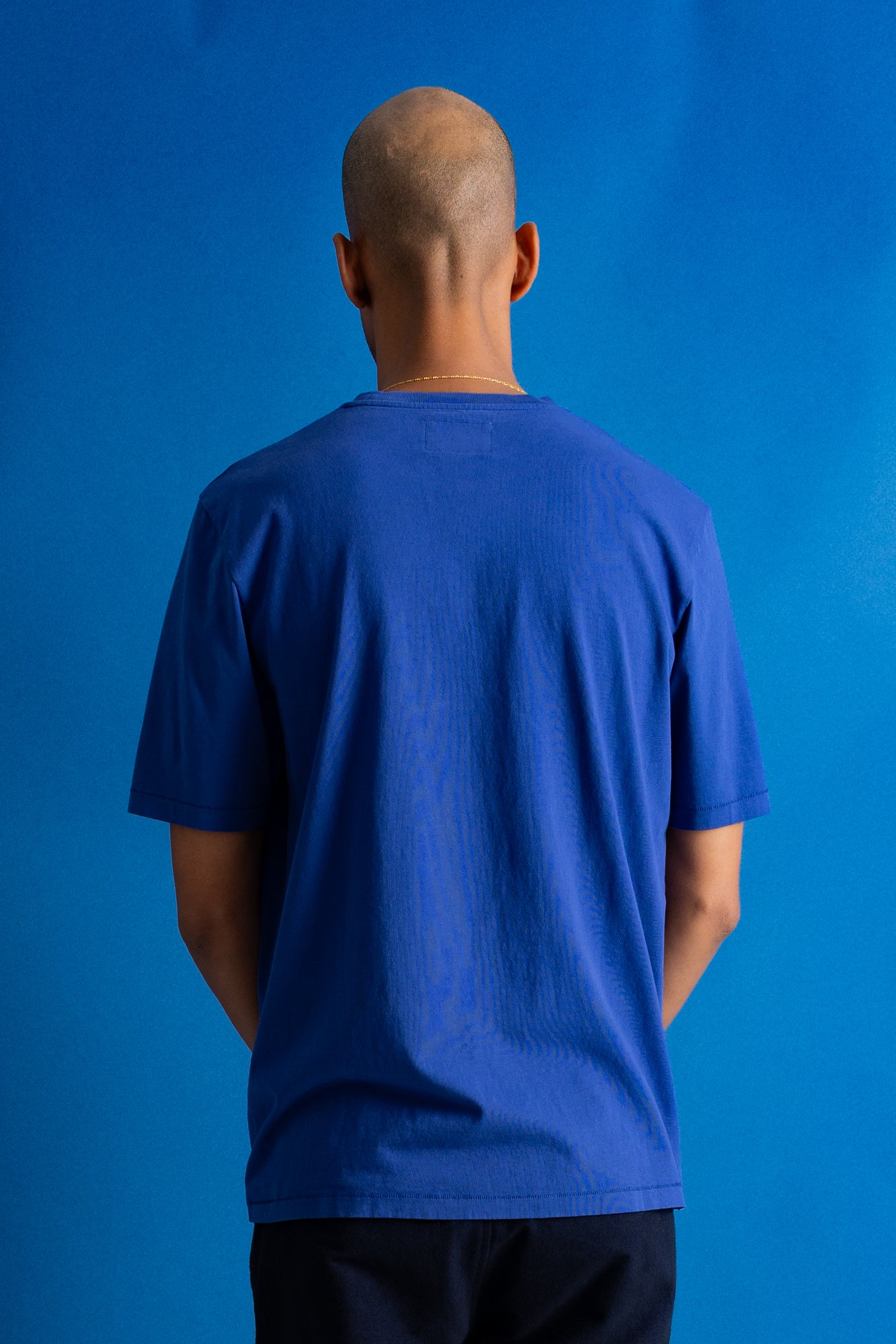 A man viewed from behind wearing a royal blue short-sleeve t-shirt and dark pants, standing against a solid blue studio background.