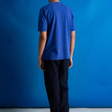 A man viewed from behind wearing a royal blue short-sleeve t-shirt and dark navy pants, standing against a solid blue studio backdrop.
