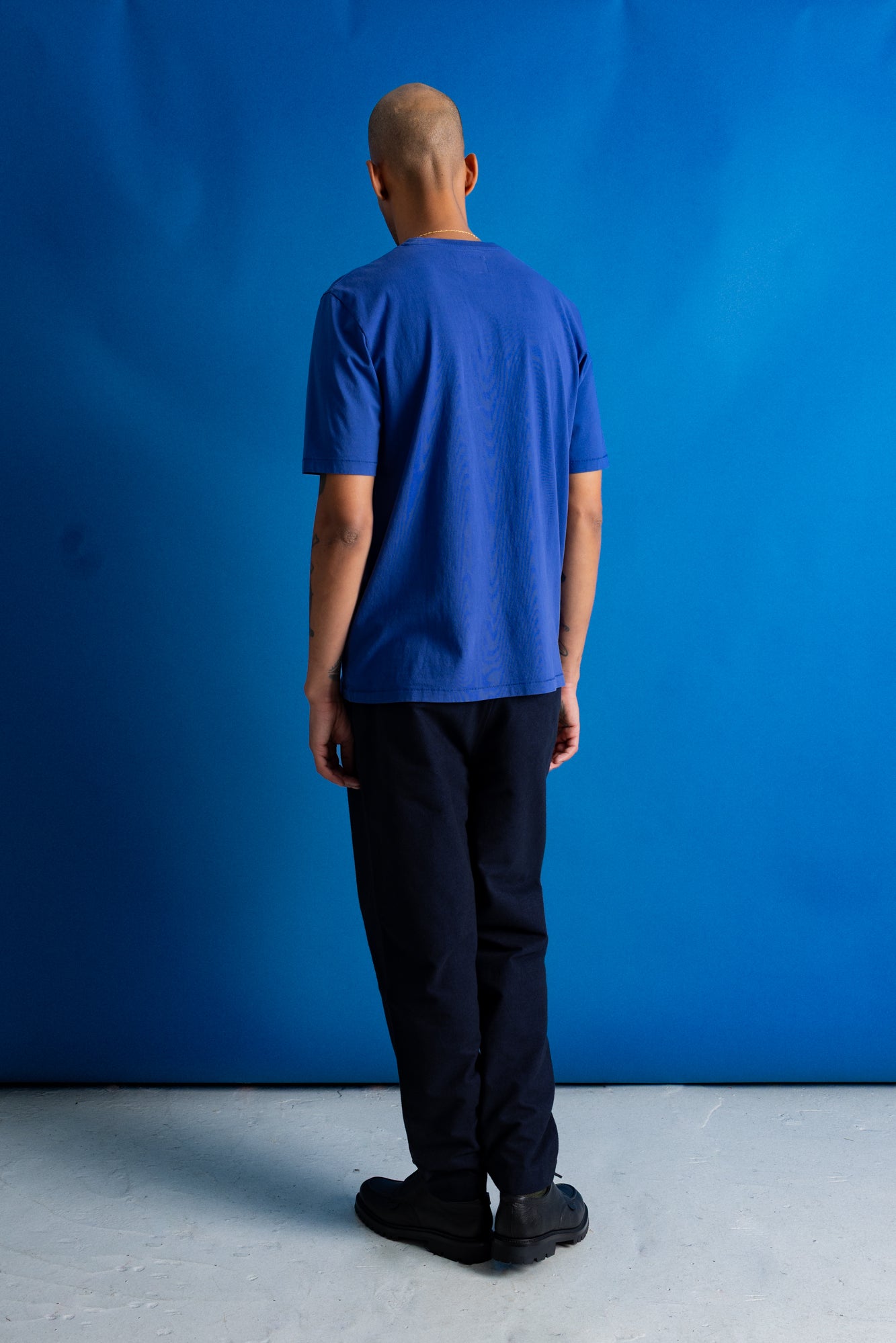 A man viewed from behind wearing a royal blue short-sleeve t-shirt and dark navy pants, standing against a solid blue studio backdrop.